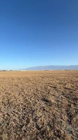 The video pans across a vast, dry, grassy field under a clear blue sky. In the distance, a mountain range is visible on the horizon. The terrain appears flat and open with sparse vegetation. Some structures, possibly buildings or distant vehicles, are scattered throughout the landscape. The overall impression is of a wide-open, somewhat arid environment.