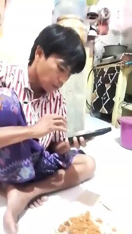 A man sits on the floor, cross-legged, holding a dark rectangular object in his hands. He is wearing a red and white striped shirt and a purple sarong with a patterned design. In front of him on the floor is a plate with food on it. The background shows a cluttered room with a water dispenser, shelves with items, and a stove. The lighting is dim.