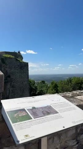 A stone wall of a castle ruin stands on the left of the frame under a bright blue sky with a few scattered clouds. In the distance, a vast landscape of green trees and fields stretches out under the wide sky. In the foreground, a white informational plaque with German text and images is displayed on a stone surface. The plaque appears to be an exhibit detailing the history or features of the castle. A person is visible sitting on the edge of the castle wall, overlooking the landscape. The sun is shining brightly, creating a clear and sunny day.