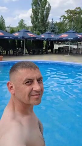 A man is in a swimming pool, taking a selfie. He has short, buzzed hair and is shirtless. The pool is a bright blue color. In the background, there is a building with a dark wooden exterior and several umbrellas with the logo URBAN CRAFT on them. There are also some trees and plants visible. The sky is cloudy.