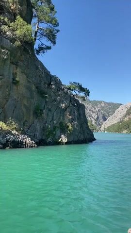 The video shows a scenic view from a boat moving on a large body of turquoise water, likely a lake or reservoir within a canyon. Steep, rocky mountains, partially covered with lush green pine trees, flank both sides of the water. The camera is positioned low, just above the water's surface, and slowly pans to the right, revealing more of the majestic canyon landscape. The sky is clear and bright blue, indicating sunny weather. The vibrant color of the water is particularly striking. The scene conveys a sense of natural beauty and tranquility, suggesting a boat tour or recreational activity in a picturesque gorge. There are no people visible, and no text is present in the frame.