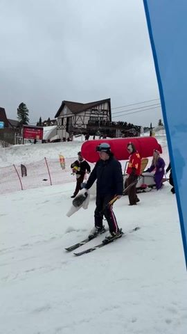 The video shows a group of people on a snowy hill. A skier is in the foreground, holding poles, dressed in a dark jacket and red-striped pants. Behind them, a homemade contraption resembling a small airplane is being pulled by people in costumes. One person is dressed as a chipmunk with a red cape and another as a mouse in a purple jumpsuit. A woman in a black coat walks alongside. The airplane-like structure has a red covering, a white body, and what appear to be skis instead of wheels. In the background, there's a building labeled "ROOKS" and a banner. The sky is overcast, and the scene appears to be a festive or humorous event at a ski resort.