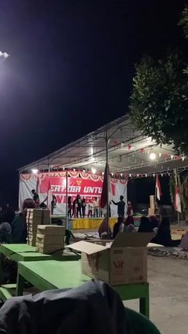 A nighttime outdoor gathering is taking place under a covered stage. A banner on the stage reads "BERTARUNG UNTUK INDONESIA" which translates to "FIGHT FOR INDONESIA". A group of children are on stage, possibly performing. Several adults are present, some watching the performance and others involved in organizing. Green tables and chairs are set up in the foreground, with boxes of what appear to be food or supplies stacked on them. The scene suggests a community event, possibly a celebration or a local gathering.