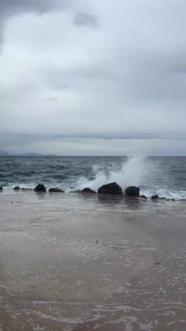 The video shows a grey, overcast day at the coast. A concrete pier or walkway is in the foreground, partially covered in water with small air bubbles. Waves crash against a series of dark rocks aligned just beyond the pier.  The sea is dark with whitecaps, and land is visible in the distant background under the cloudy sky.
