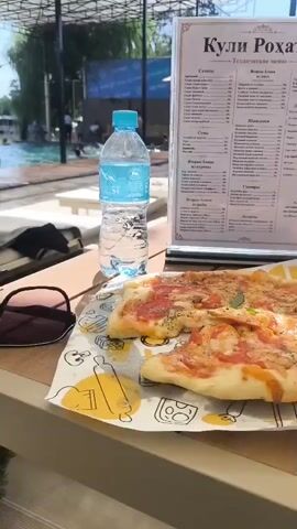 The video begins with a close-up of a pizza and a bottle of water on a table. A menu is visible in the background. The camera then pans to show a man in swim trunks bending down near a green bush, with a swimming pool and people in the background. The scene shifts to show a view of a pool area with several people swimming and relaxing. A man is seen walking past the camera towards the pool. Another shot shows a wider view of the pool area with more people enjoying the water. The video ends with a shot of the pool area, with people swimming and a woman standing near the edge of the pool. The environment appears to be a resort or a water park on a sunny day.