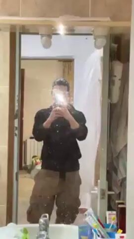 A person with short, dark hair takes a selfie in a bathroom mirror. The person is wearing a dark, button-up shirt with the sleeves rolled up and khaki cargo pants. They are holding a light blue iPhone, and the flash creates a bright glare that obscures their face. The mirror is dirty with many water spots. The bathroom features a sink with a variety of items on it, including toothbrushes, toothpaste, and other toiletries. Behind the person and visible in the mirror is a doorway leading to another room under renovation. There is a radiator, some construction materials, and a white plastic sheet visible through the doorway. A white towel hangs on a rack to the right of the mirror.
