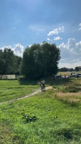 A person is riding a bicycle on a dirt path through a grassy field. In the background, there is a large tree and a parking lot with several cars. The sky is blue with white clouds, and the sun is shining. The overall scene appears to be a rural or park-like setting on a sunny day.