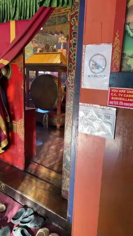 From the threshold of an ornate doorway, the camera peers into a monastery.  The doorway is flanked by an arrangement of red, green, and gold drapes.  To the right is a red doorframe with signs posted reading, "NO PHOTO" and "PLEASE TAKE OFF YOUR SHOES & SLIPPERS". The doorframe itself is elaborately decorated.  Inside the room, several monks dressed in maroon robes sit on cushions along a wall.  There is a large drum with a gold canopy.  A monk walks toward the doorway, his robe partially covering his face.  The inside of the room is ornately decorated with gilded figures and patterns.
