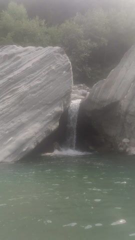 This media file depicts a natural scene at the Baragran, India location, likely a river or a waterfall. The primary subjects are large, grey, striated rocks that form the landscape, with a body of greenish water in the foreground. A small waterfall or rapid is visible cascading between the rocks. Dense, green foliage can be seen in the background, suggesting a wooded or mountainous area. The lighting indicates it is daytime, possibly morning or late afternoon, with a hazy or misty quality to the air, and bright sunlight filtering through from the left side of the frame. The overall mood is serene and natural, emphasizing the raw beauty of the environment. There is no visible text in the media file.