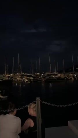 The video shows a nighttime scene at a marina in Kumbor, Montenegro. In the foreground, two people are seated, viewed from behind. Their faces are not visible, and their attire is casual. A metal chain fence separates them from the water. The background is dominated by a dark, cloudy night sky and a harbor filled with numerous sailboats, their masts creating a striking silhouette against the twilight. Lights from the boats and possibly from the shore are visible, reflecting on the calm water. The overall mood is tranquil and serene, suggesting a peaceful evening by the sea. The time of day is clearly night, with a touch of drama provided by the cloud formations. No text is visible in the video.