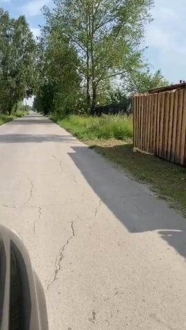 The video shows a first-person perspective of driving down a paved road lined with trees and grass. The road is cracked in places. On the right side of the road, there is a wooden fence and a small building visible. The sky is partly cloudy. The footage appears to be taken on a sunny day.