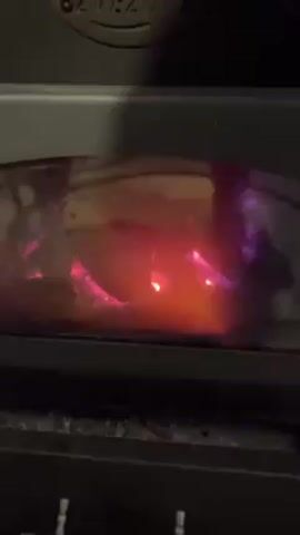 The video shows a close-up of a burning fire inside a black wood-burning stove. The fire glows orange and pink behind a glass door. The word "ZVEZDA" is visible on the top of the stove. A metal handle is below the glass door. The scene appears dimly lit, emphasizing the fire's glow.