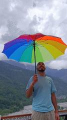 Young man holds rainbow umbrella outdoors, smiles skyward