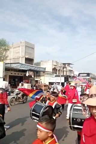 A street parade is underway. A group of young people in matching red and black uniforms, some carrying drums and others holding flags, are marching. A woman in a red hijab and a straw hat is visible in the foreground, looking towards the parade. In the background, buildings line the street, and a large bus with decorations is part of the procession. The sky is cloudy. The overall atmosphere appears festive.
