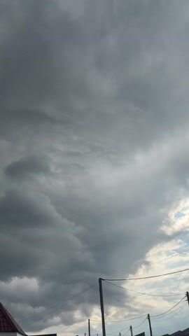 The video captures a view of a cloudy sky over what appears to be a residential area. In the lower portion of the frame, the corner of a house with a dark red roof is visible on the left. Further to the right, a section of a wall with a window and a light fixture is seen. The ground in front of the house is covered with small gray stones and patches of grass. Power lines and utility poles are also present in the lower right part of the image, suggesting an outdoor setting. The sky is predominantly filled with dark, heavy clouds, indicating overcast weather, possibly a storm is approaching or passing. The overall impression is of a somber and potentially windy or rainy day.