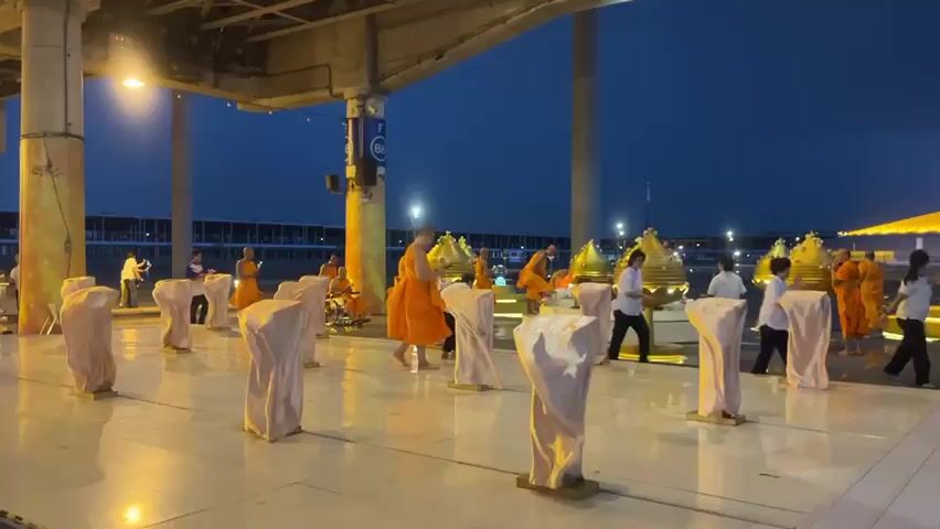 The video shows a group of Buddhist monks in orange robes gathered in an open area at dusk. They are standing in front of a series of golden, urn-like structures arranged in a semi-circle. Some monks are walking, while others are stationary, seemingly engaged in prayer or ritual. In the background, a large golden dome structure is visible, illuminated by lights. Several white, fabric-covered pillars or pedestals are positioned in the foreground, partially obscuring the view of the monks and the golden structures. The sky is a deep blue, indicating twilight. The ground is paved and reflective, suggesting it may be wet. Some individuals in white clothing are also present, interacting with the monks or observing the scene. One person is seen in a wheelchair. The overall atmosphere appears serene and spiritual.