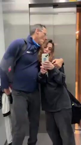 The video begins with a selfie taken by a woman in an elevator with a man. The woman wears a black sweatshirt and trousers, and the man wears a blue jacket, black trousers, and glasses. He has a backpack on and is holding a white jacket. The elevator door opens, revealing a beige hallway with brick-like walls. The camera moves quickly, then focuses on a grey door with a black handle, labeled with a yellow sign that reads "SUITE 2207-2277."