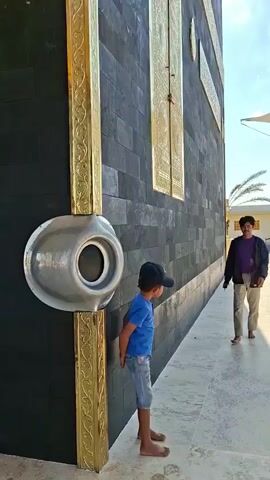 Two males, one an adult and the other a child, stand beside a large black structure with gold accents. The child, wearing a blue t-shirt and denim shorts, leans against the structure, barefoot. The adult, wearing a purple shirt and beige pants, walks towards the child and places a hand on the child's head. The adult then appears to touch or examine the structure. The environment appears to be outdoors, with a tiled walkway and a blue sky with scattered clouds. A white canopy-like structure is visible in the background, along with a building and some palm trees.