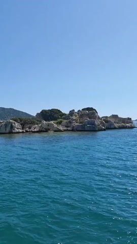 The video captures a scenic boat journey through calm, vibrant turquoise waters under a pristine, cloudless blue sky. The perspective is from a moving boat, with a chrome railing briefly visible in the foreground at the beginning of the video (0:01-0:02).

On either side of the boat's path and in the distance, rugged, rocky coastlines and small islands are visible. These landforms are characterized by light-colored rock formations and sparse green vegetation, including bushes and small trees. Larger, verdant hills form the backdrop in several shots.

The activity depicted is a serene passage through a beautiful coastal environment, likely during a boat tour or transfer. The bright sunlight, clear sky, and calm water indicate a perfect sunny day, possibly in the late morning or early afternoon. The landscape aligns with coastal areas found in Demre, Turkey. No people are explicitly visible, and there is no discernible text within the video frames.