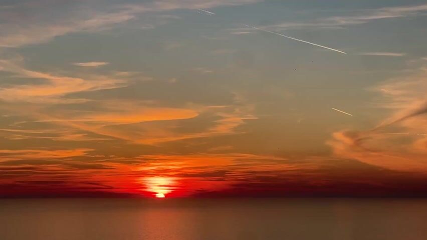 The video shows a sunset over the ocean, with a fiery orange sun partially visible on the horizon. The sky is a mix of blues, oranges, and reds, with streaks of clouds and contrails from airplanes. The sea appears calm and dark, reflecting the colors of the sky. The sun is the focal point, shining brightly against the dark sea.