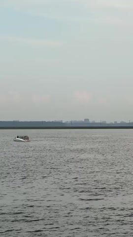 A wide shot shows a large body of water with a distant city skyline on the horizon. In the foreground, a small white boat with a red stripe travels across the water. The sky is overcast and a muted gray. The water is choppy, with small waves.