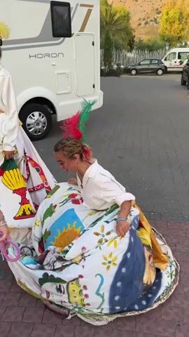 A young woman with pink and green feathers in her hair, wearing a white shirt and a flowing skirt decorated with colorful painted images of cacti, a sun, and faces, is kneeling on a paved surface in front of a white RV. She is holding the edge of the skirt, which is spread out around her like a large circle. Another person, wearing a white t-shirt and dark shorts, is partially visible on the left, also holding onto the skirt. The background shows a paved parking lot with several cars and a white van, and beyond that, a hillside with some greenery. The lighting suggests it is daytime.