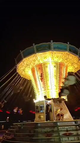 At night, a carnival swing ride is in operation. The ride is brightly lit with yellow and orange lights, illuminating the central pillar and the rotating arms with attached swings. Several people are visible on the swings, their bodies blurred due to the motion. A small booth is located at the base of the ride, with at least two people standing inside and two people sitting on steps in front of it. Other figures are visible in the background, suggesting a crowded environment. The ground beneath the ride is dark, and there are some faint lights visible in the distance. The overall impression is of a lively amusement park at night.