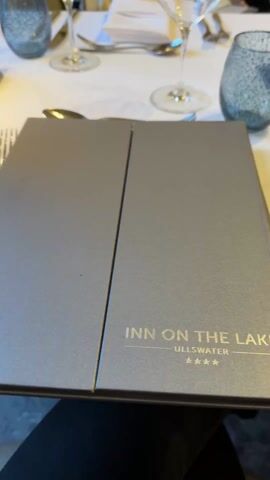 The video shows a table in a restaurant with a view of a rainy landscape. The table is set with cutlery, glassware, and a menu from a place called “Inn on the Lake”. The camera pans to a window revealing an outdoor patio area with tables and chairs, all wet from the rain. In the distance, there are mountains shrouded in mist, along with a gazebo near a body of water, likely a lake. The overall environment suggests a serene, though wet, dining experience.