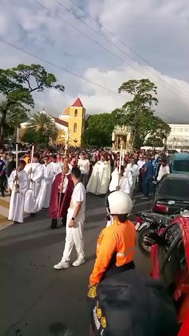 This is a vertical, outdoor video taken during daylight. The scene appears to be a religious procession in a street. In the foreground, three people are partially visible from behind, wearing orange and blue safety vests, and one person is holding a smartphone. In the middle ground, a procession of people dressed in white robes are walking, some carrying candles and a large cross. Behind them, a larger crowd of onlookers is visible. In the background, a yellow church building with a red roof stands against a cloudy sky. Trees and power lines are also present in the background. The overall atmosphere seems festive or solemn, consistent with a religious event.