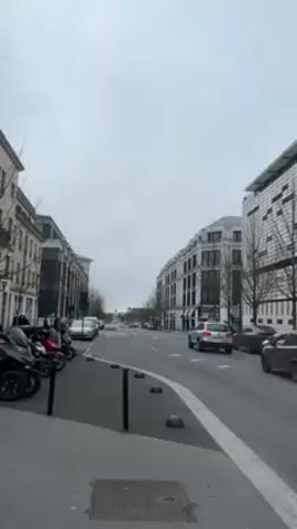 The video shows a street scene, likely in Europe, judging by the architecture and vehicle models. The street is lined with multi-story buildings, some of which have classic architectural details. It appears to be a cloudy, overcast day. Several cars are moving on the street, and a few are parked along the side. There are also a couple of three-wheeled motor scooters parked on the sidewalk. There are a few bare trees visible. A traffic light in the distance is red, and the other lights show green. The street is marked with a white line dividing traffic lanes.