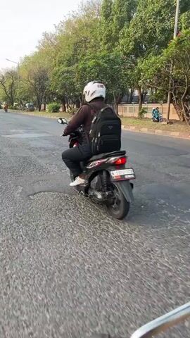 The video shows a person riding a motorcycle on a paved road. The rider is wearing a white helmet and a dark jacket, and they have a black backpack on. The motorcycle is black and silver, with a visible license plate. The road is lined with trees, and there are buildings in the background. The sky appears to be clear. The camera seems to be mounted on another vehicle, following the motorcycle from behind and slightly to the side. The overall scene suggests an outdoor, daytime environment, likely in an urban or suburban setting.