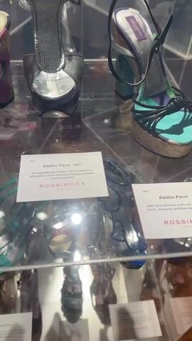 This video shows a display of women's shoes in a museum or gallery setting. The shoes are presented in glass display cases. The primary subjects are the shoes themselves, which appear to be designer items, with some labels visible. The scene is indoors, with wooden flooring and white walls. Paintings hang on the walls in the background.

In the foreground, several pairs of high-heeled shoes are visible, including sandals with colorful straps and wedge heels. One label reads "Emilio Pucci 2007 karung laminato argento, tacco e plateau in plexiglass / silver laminated karung, plexiglass heel and plateau." Another label is partially visible with the text "ROSSIMODA YEARS 1931-2007". Further in the background, a young woman with blonde hair is seen from behind, wearing a white ribbed tank top and a black skirt. She is looking at the display.

The lighting suggests it is daytime, likely indoors with artificial lighting supplementing natural light, as evidenced by reflections on the glass. The overall atmosphere is one of an exhibition or curated display of fashion. The mention of "ROSSIMODA" and "Emilio Pucci" suggests a focus on high-end fashion and possibly a retrospective of this designer or brand. The location context of Stra, Italy, indicates this exhibition is taking place in that region.