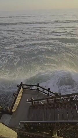 The video shows a wooden boardwalk or staircase leading down towards the ocean. Waves are crashing against the shore, creating sea spray that rises up. Several people are visible on the boardwalk, some of whom appear to be taking pictures or enjoying the view. The sun is low in the sky, casting a warm glow on the scene, suggesting either sunrise or sunset. The coastline features green vegetation on a cliff face.