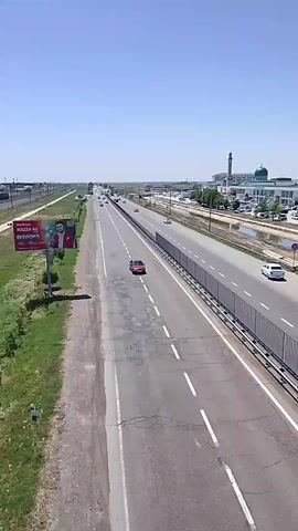 The video is taken from an elevated perspective, possibly a bridge, overlooking a multi-lane highway under a clear, blue sky. The highway is relatively straight, with a median barrier running down the center. Cars are seen moving in both directions. To the left of the highway, there is a railway line and a large billboard featuring a man in a red suit and text in a non-English language. The billboard has the words "MAZZA 60" in a prominent font. On the right side of the highway, there is a complex of buildings including a mosque with a green dome. The area surrounding the highway is green with vegetation. The general impression is of a sunny day in a suburban or semi-urban environment with moderate traffic.