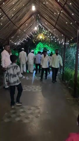 A group of people, mostly men, are walking through a covered walkway decorated with string lights. The walkway has a thatched roof. A child in a red shirt is in the foreground and partially obscures the view. Most of the men are wearing white shirts. One man is wearing a light pink shirt and tan pants. One boy is wearing a black and white patterned shirt. There are plants on either side of the walkway. The ground is paved with what appears to be concrete. The lighting is dim.