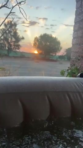 The video shows a time-lapse of a sunset viewed from behind an inflatable object. The sun is visible in the center of the frame, with rays of light emanating from it. Clouds are scattered across the sky, some tinged with orange and pink hues. In the foreground, the inflatable object takes up the bottom portion of the screen, appearing tan and textured. Behind the inflatable, a blurred field is visible, with a line of trees in the distance. The overall scene conveys a sense of calm and natural beauty.