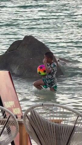 A young boy stands in shallow, clear ocean water, holding a colorful beach ball. He is wearing a patterned t-shirt and green shorts. Behind him, a large, dark rock juts out of the water. In the foreground, a portion of a sign with the words "HAPPY HOUR" in red and white text is visible, along with two white woven chairs. The water is calm with small ripples. The lighting suggests it could be late afternoon or early evening. The scene appears to be a beach resort or a similar coastal area in Ko Tao Subdistrict, Thailand. The boy's posture and the presence of the ball suggest he is either about to play or has just finished playing.