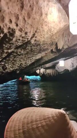 The video shows a view from a boat within a cave. The water is dark and reflective, with other boats visible in the distance, illuminated by a light. The cave ceiling is low and textured, with visible stalactites. The foreground features a traditional Asian conical hat.