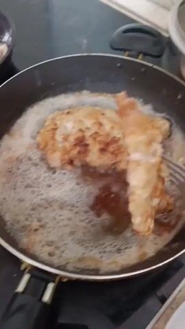 This media file captures a close-up view of food being prepared in a kitchen. The primary focus is on several pieces of breaded and fried meat, likely chicken or pork cutlets, sizzling in a black frying pan filled with bubbling oil. A fork is used to lift one of the cutlets, revealing its golden-brown, crispy exterior. In the background, a pot with a lid is visible, possibly containing pasta or another side dish. A partially visible bottle of cooking oil and a floral-patterned cloth suggest a home kitchen environment. The lighting appears to be artificial, indicating the scene could be taking place at any time of day. There is no visible text within the media file. The activity is the frying and plating of meat cutlets. The setting is a kitchen, and the location context is Penza, Russia.