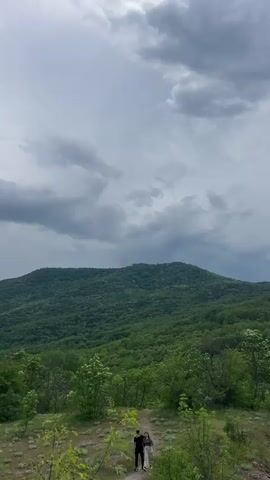 The video shows a landscape with a large green mountain in the background, partially obscured by a cloudy sky. In the foreground, a trail is visible, where a couple is walking away from the camera. The vegetation surrounding the trail includes low bushes and trees with white blossoms. The overall setting suggests a rural or natural environment.