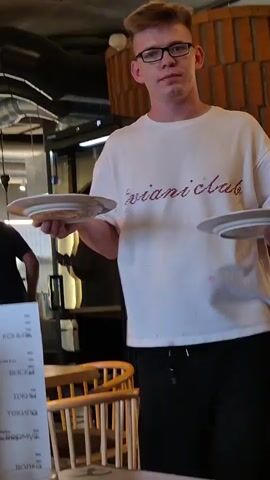 A man wearing a white t-shirt and black pants, with glasses and a shaved head, stands in a restaurant. He is holding a plate with a piece of food on it, possibly a pastry or bread with toppings. To his left, another person is visible in the background. A menu is propped up on the table in front of him, with a glass of liquid next to it. The man places the plate on the table and then reaches for a chair. The restaurant appears to be casually decorated with wooden furniture and exposed brick walls.