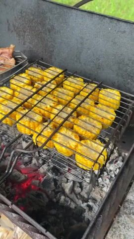 This is a close-up, overhead shot of corn on the cob and meat being grilled over hot coals. The corn is cut into rounds and arranged in a grilling basket, with visible grill marks forming on the bright yellow kernels. Next to the corn, in a separate grill basket, is raw meat, appearing to be ribs or strips, with some red onion pieces interspersed. Further back and slightly higher, another grill basket contains what look like marinated chicken wings, seasoned with a reddish spice mix. The scene is set outdoors, with green foliage visible in the blurred background, suggesting a natural or garden setting. The hot coals are glowing red, indicating the grilling is in progress. The lighting suggests it is daytime, likely a sunny or overcast day given the bright colors and lack of strong shadows. The overall impression is one of a casual outdoor barbecue or cookout. No people are visible in the frame. There is no visible text. The context provided indicates the location is Levkovo, Russia.