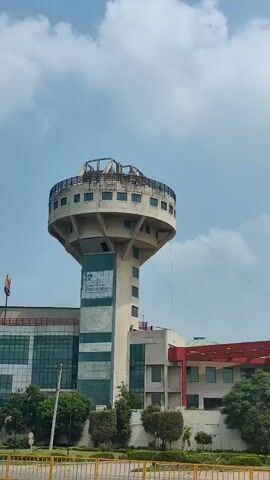 The video shows a tall, round water tower with a circular observation deck at the top. The tower is a light beige color and has a cylindrical base that supports the wider top section. Several windows are visible around the observation deck. A modern building with large glass windows is situated to the left of the water tower. Part of the building is under construction. A yellow barrier is visible in the foreground, and a road runs in front of the building and tower. A white SUV is driving on the road, and a motorcycle with a rider is also passing by. The sky is partly cloudy.