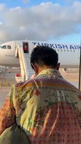 A short video captures a sunny outdoor scene at an airport. Two men walk towards the steps of a Turkish Airlines plane, preparing to board. The man in the foreground wears a patterned shirt and carries a grey neck pillow. The man behind him wears a dark shirt and carries a dark bag. The white plane with "TURKISH AIRLINES" written on its side is visible, with a ground crew member in an orange vest standing at the top of the stairs. Airport ground equipment is also visible in the background, including a white "SCHOPF" vehicle with "ECE" on its side. The sky is bright blue with scattered white clouds. The video cuts to a different angle, showing more airport vehicles and a water tower-like structure in the distance under the same blue sky.