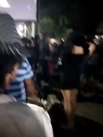 The video opens with a blurry shot of a large outdoor screen displaying an image of people, illuminated by bright lights. The screen is positioned behind a crowd of individuals, mostly out of focus, who are gathered in front of it. Some people are raising their hands. The scene appears to be a nighttime event.

The camera then cuts to a woman in a black dress standing in what appears to be a street or open area. She has long, dark hair and is striking a pose, touching her temple. Behind her, a crowd of people is visible, also in a nighttime setting. Some are blurry, but others are more distinct, including a man in a grey hooded jacket.

The video returns to the outdoor screen, showing a similar scene of a crowd watching something projected. The lighting is still bright on the screen, and the crowd in front of it is mostly indistinct. People are again seen with their arms raised. The overall impression is of an outdoor gathering or event.