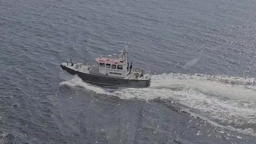 A pilot boat is moving through the water, leaving a white wake. The boat is dark with a white superstructure and has the word PILOT written on it in red. There are several people visible on the deck of the boat. The water is choppy and appears to be a deep blue color. The camera is positioned at an angle that suggests it is also on a boat or a higher vantage point.