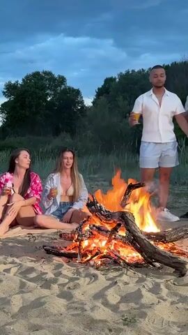 A group of four people are standing and sitting near a bonfire on a sandy beach. Two women are sitting on a beach blanket and holding drinks, while two men are standing. The man on the right is wearing ripped jeans and a patterned button-down shirt, and the other man is wearing light blue shorts and a white button-down shirt. They are all facing the bonfire. The sky is overcast, and there are trees in the background.
