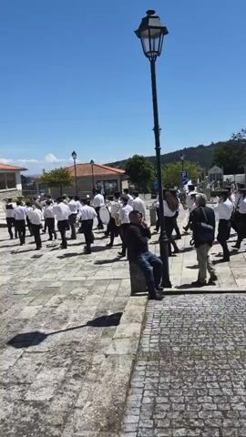 A marching band in white shirts and black pants performs on a stone paved square under a clear blue sky. A man sits casually on a stone structure next to a lamppost, seemingly watching the band. There are other people walking around the square, and buildings with orange roofs in the background.