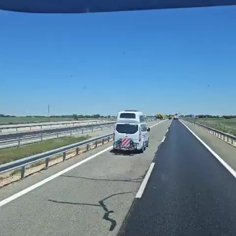 A video shows a driver's perspective on a highway, under a clear blue sky. A white camper van drives in the same direction. Beyond the highway barrier on the left, a large black bull statue is visible. A white semi-truck with a green trailer subsequently drives in the opposite direction, passing by.
