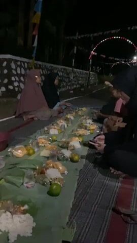 The video shows an outdoor evening gathering where a group of people are sitting on the ground around a long arrangement of food laid out on banana leaves. The food appears to be served in individual portions, wrapped in plastic, and includes rice, bread, and what looks like fruit. Several people are visible, some wearing hijabs, others in casual clothing. They are seated on mats or directly on the ground. In the background, there is a stone wall, some decorative flags, and lights. The overall atmosphere suggests a communal meal or celebration.